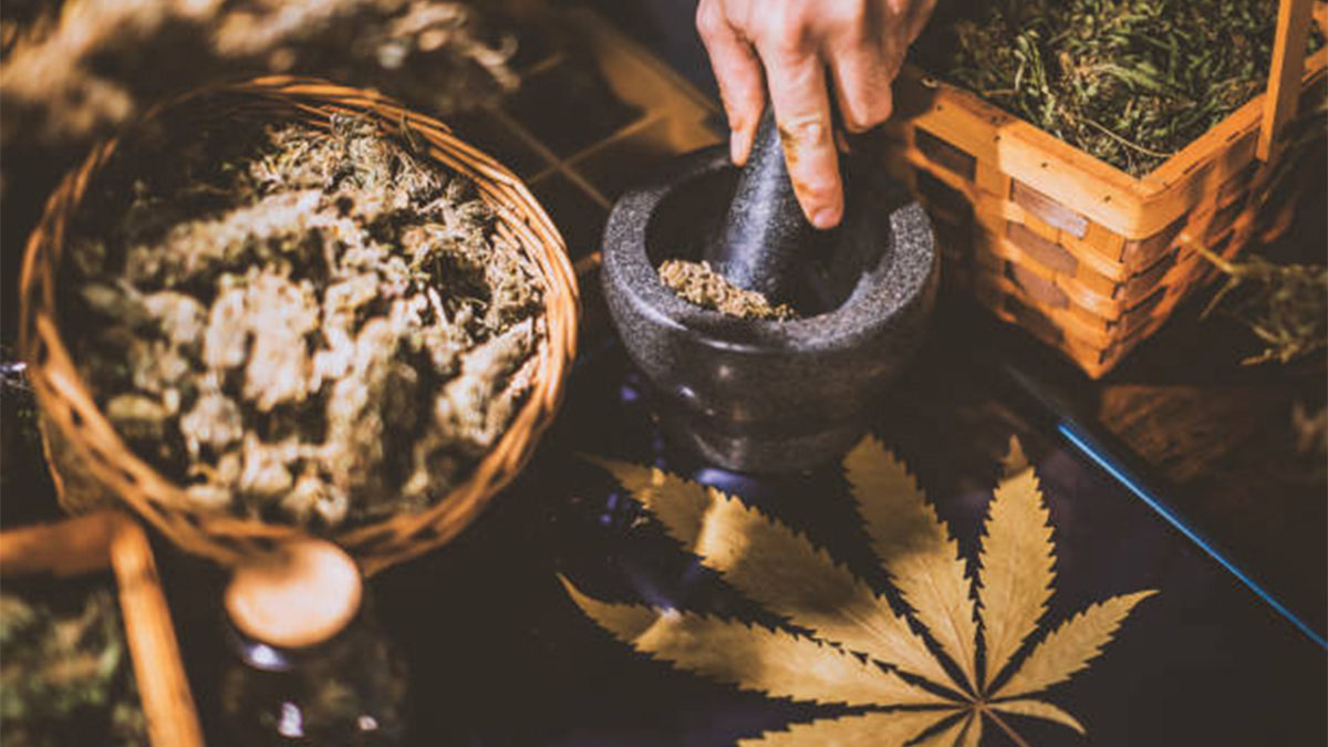 Grinding weed in a mortar and pestle with a basket of buds on the side and a marijuana leaf in front